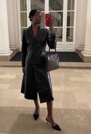 The frame bag trend is shown in a street-style photo of a woman standing in front of a mirror wearing a black leather dress with black heeled mules and a brown frame bag