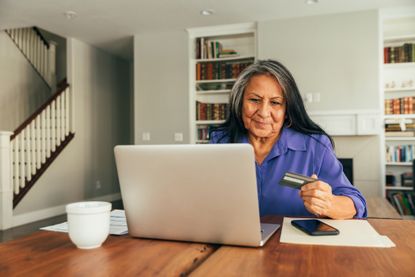 A senior adult woman sits at the dining table paying bills and banking using her mobile laptop.