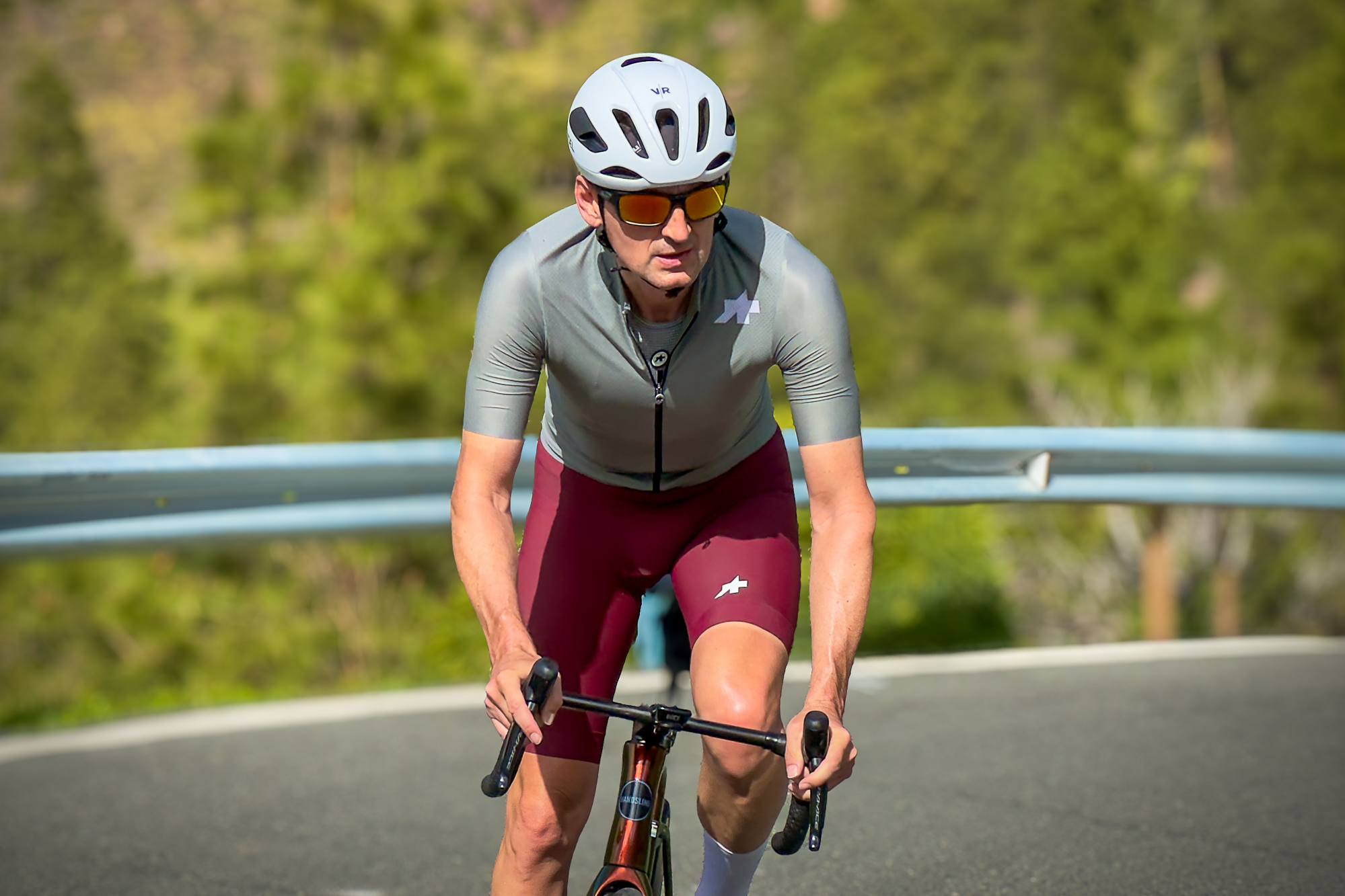 Man wearing a green jersey, red shorts and white helmet riding a road bike up a hill out of the saddle towards the camera