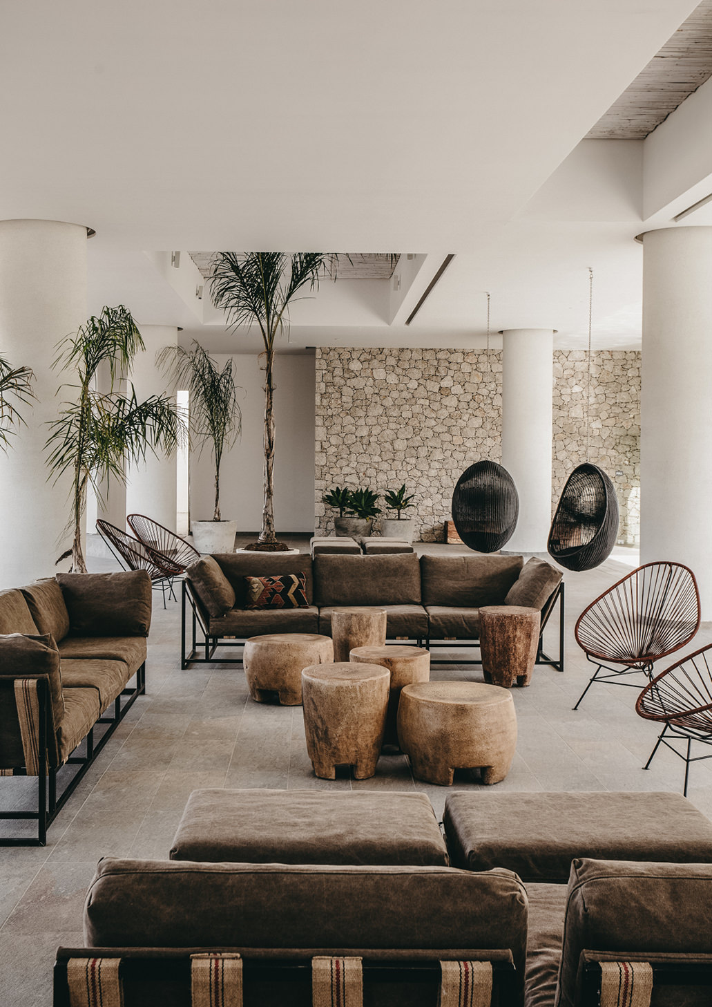 A modern rustic-decorated hotel lobby pullulates with wood, brown velvet, wrought iron, and stone-based furnishings and details in a naturally lit, plant-filled setting.
