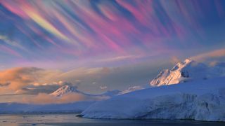 nacreous clouds appear as wispy multi-color iridescent clouds streaking across the sky above snowy mountain tops. 