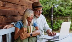 A mature couple sit at a table outside, looking at a computer.