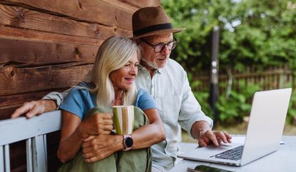 A mature couple sit at a table outside, looking at a computer.