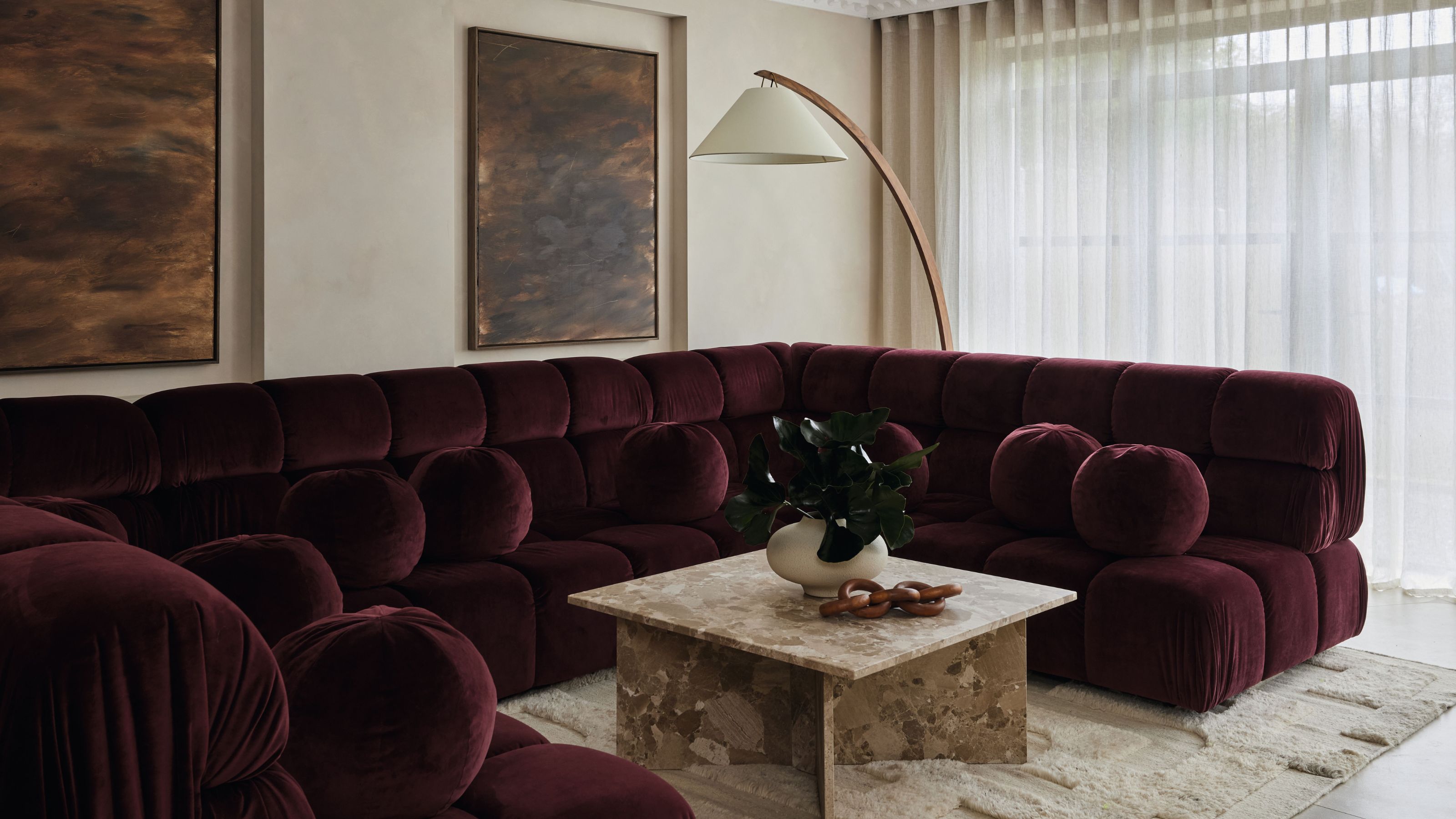 A luxurious living room designed by Amy Stoddart, featuring a deep plum velvet tufted sectional sofa and a square marble coffee table on a textured cream rug.