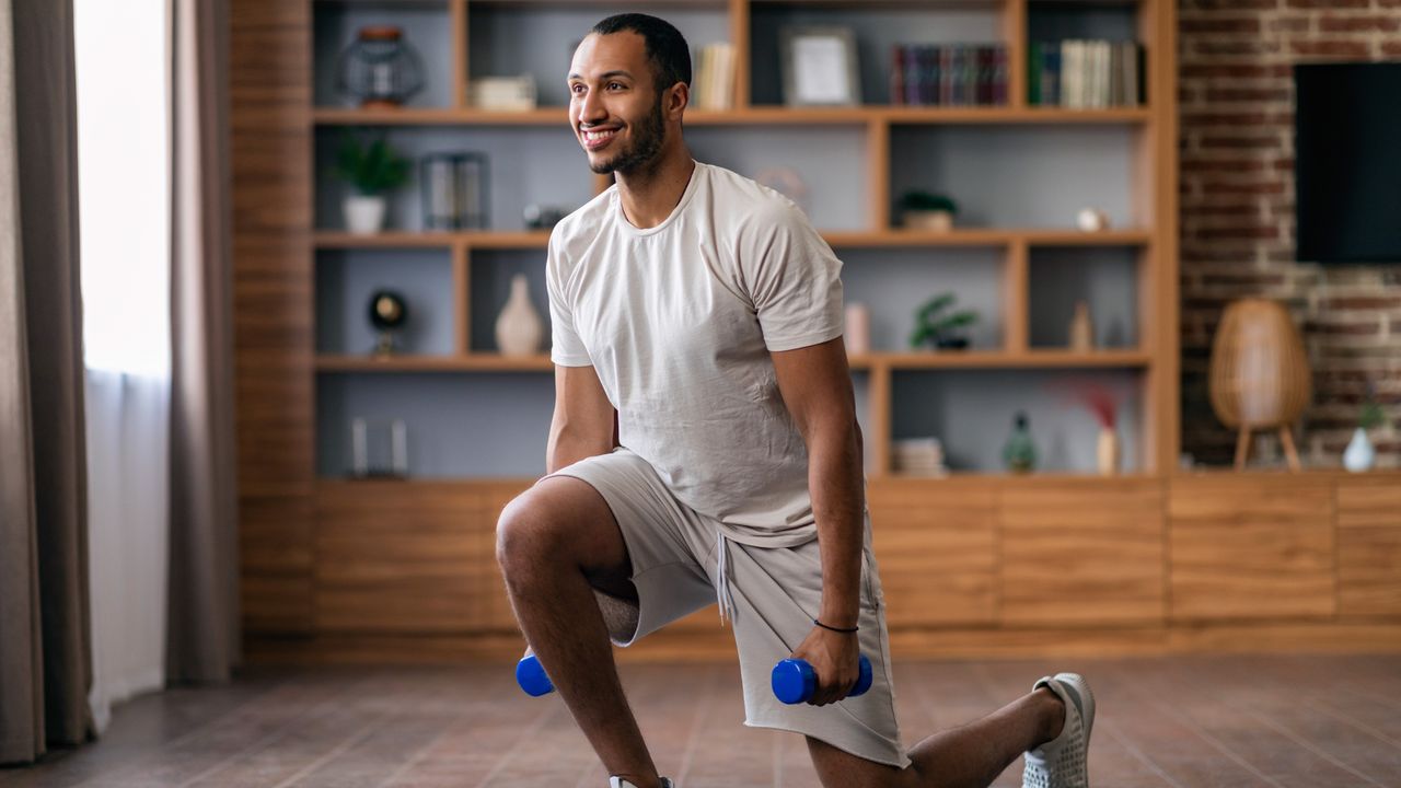 A man performs a reverse weighted lunge in his living room. He wears shorts, t-shirts and sneakers and holds a pair of dumbbells in his hands, which hang by his sides. He&#039;s in the middle of a lunge, with his left leg behind him and left knee hovering just above the floor, and his right leg bent at a right angle in front of him. Behind him we can see a bookcase filled with decorative items and a television mounted on a brick wall.