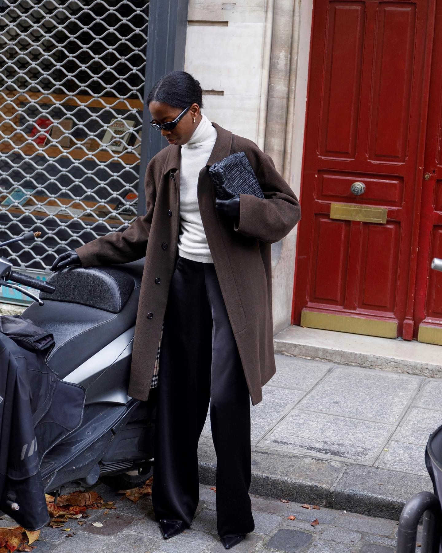 Sylvie wears a brown coat, white turtleneck, black clutch, black satin pants, and pumps.