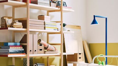 IKEA PS shelving unit decorated with books, files, and trinkets, against a yellow and white wall, with a blue floor lamp.