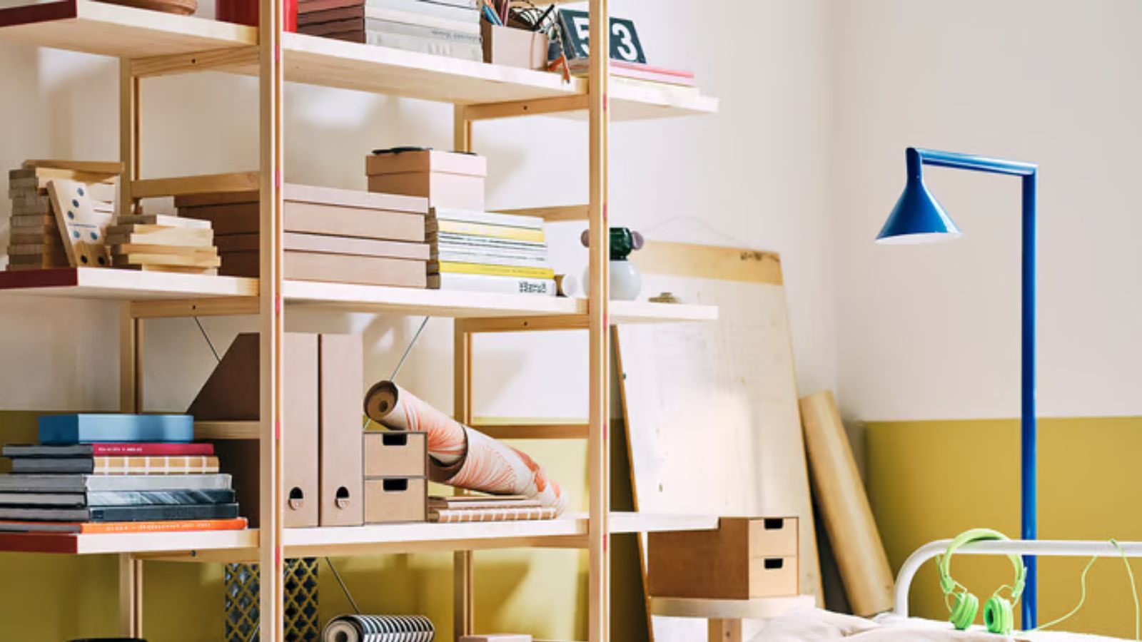 IKEA PS shelving unit decorated with books, files, and trinkets, against a yellow and white wall, with a blue floor lamp.