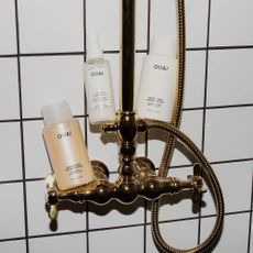 Three Ouai bottles balanced on gold shower, white tiles.