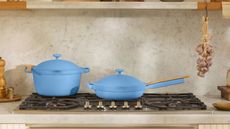 A blue Dutch oven pan, and a matching blue frying pan with ids on a black gas burning stove top. There is a matching greige stone backsplash and countertop, and garlic bulbs hand from a shelve above. 