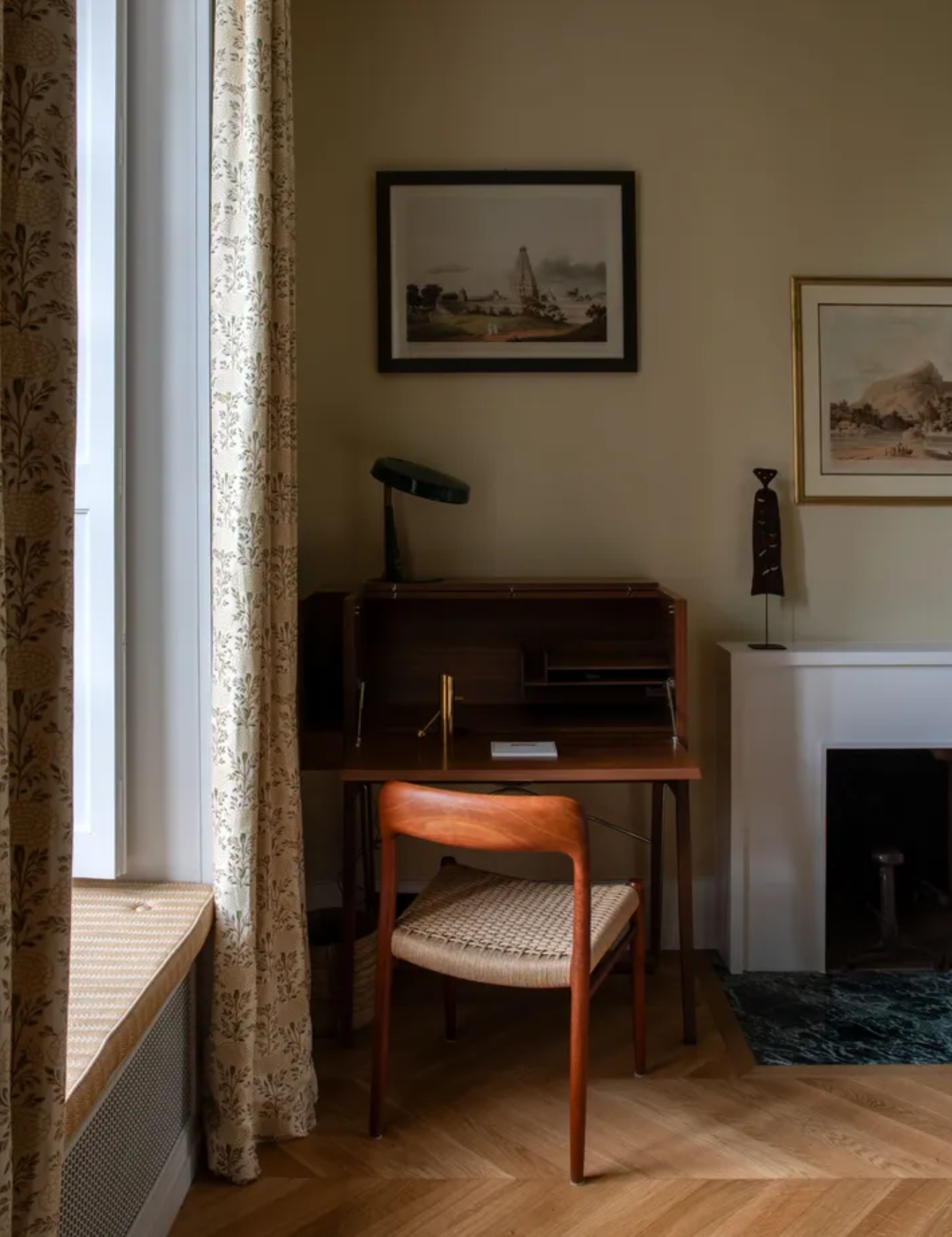 A small wooden writing desk with a matching chair in the corner of a neutral painted room with wooden herringbone flooring.