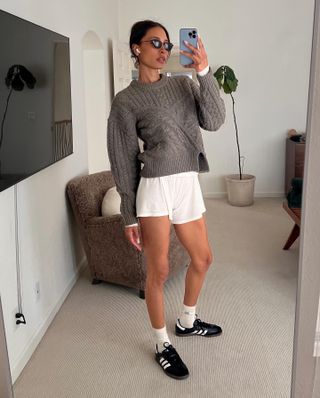 Woman wears Adidas sambas, white shorts and a chunky grey sweater taking a mirror selfie.