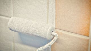 Close up of mini roller applying white paint to beige kitchen tiles