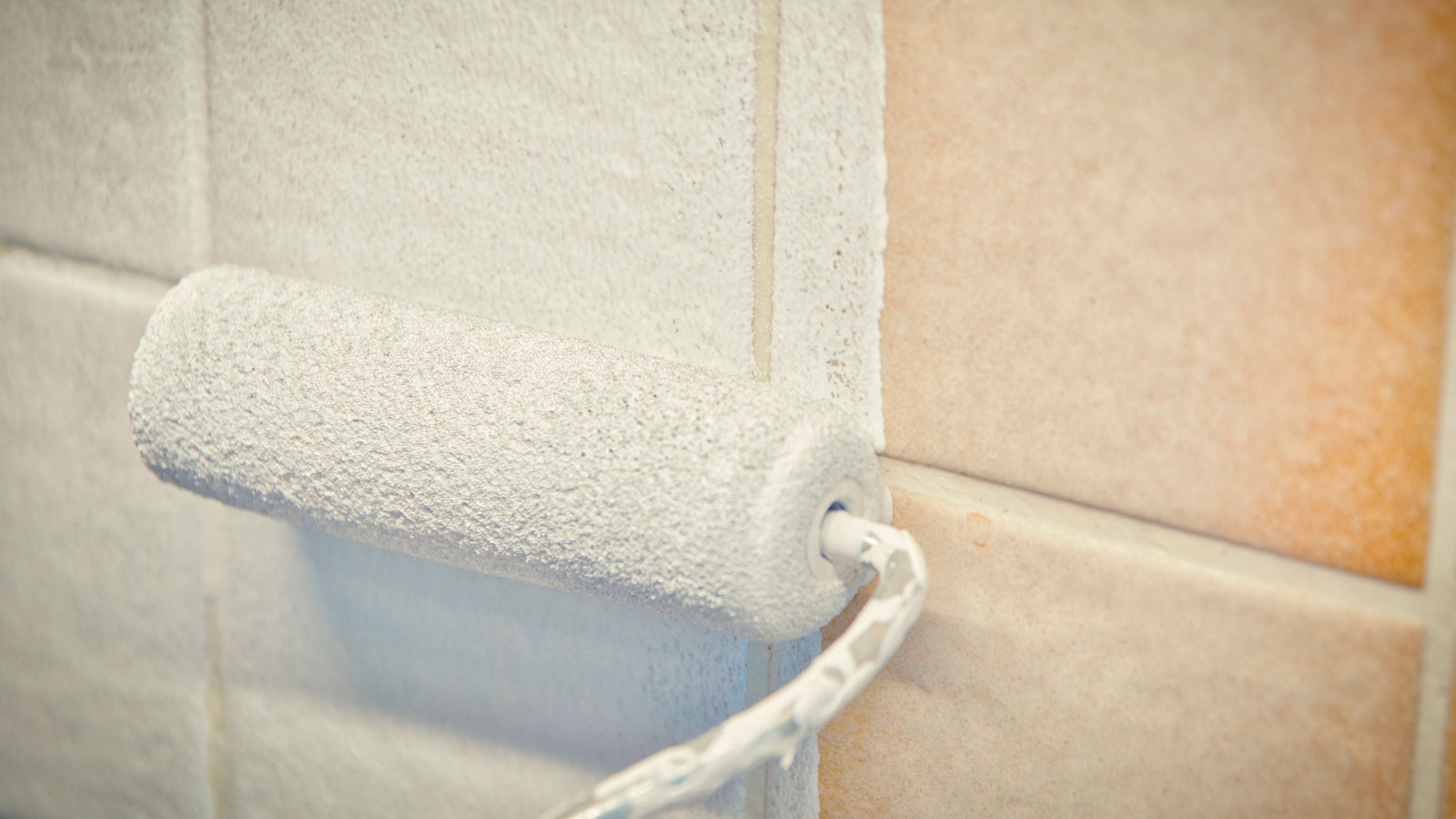 Close up of mini roller applying white paint to beige kitchen tiles