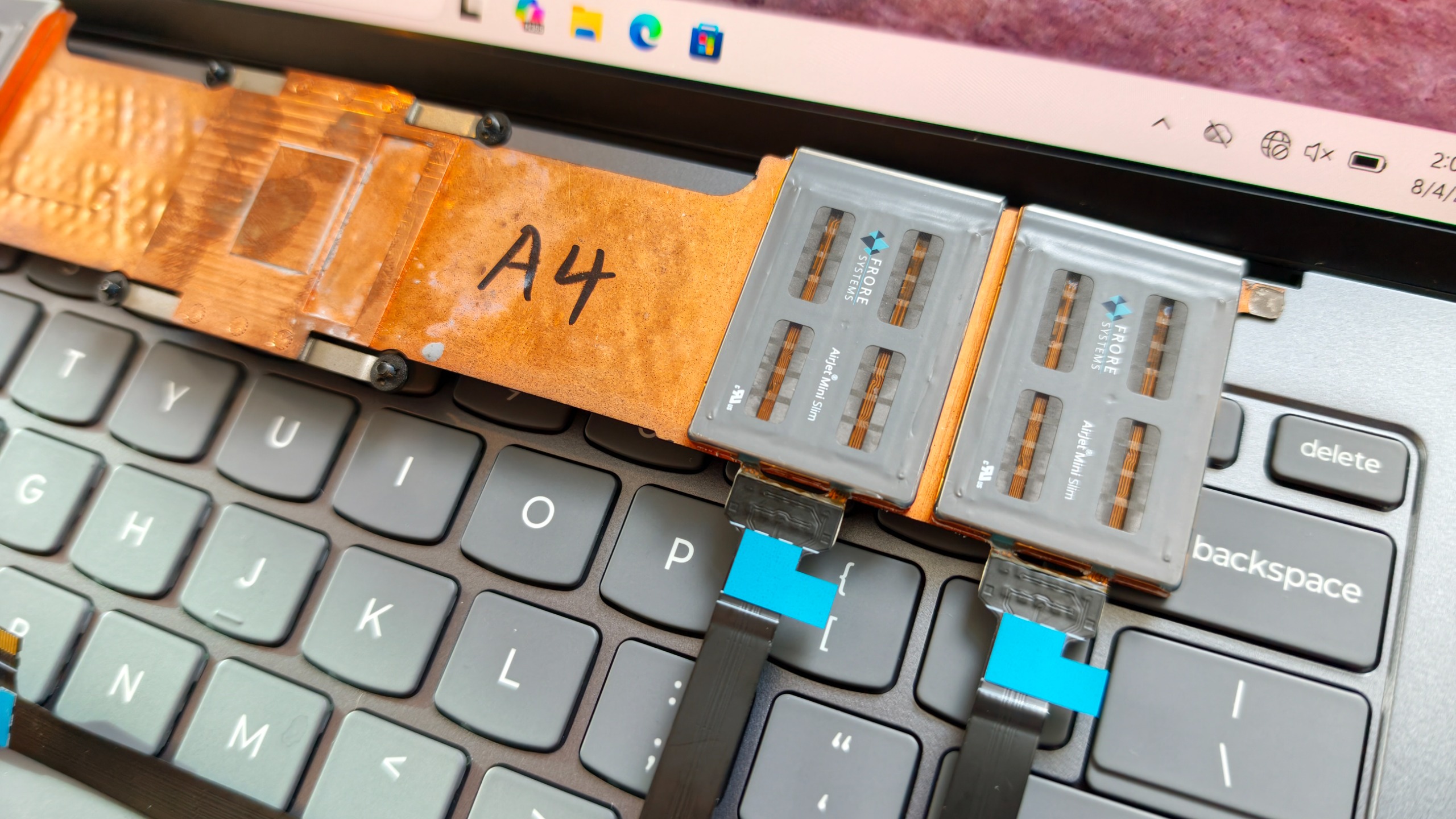 Close-up of two Frore AirJet Mini Slim solid-state cooling chips installed on a copper heat pipe strip above gray laptop keyboard keys.