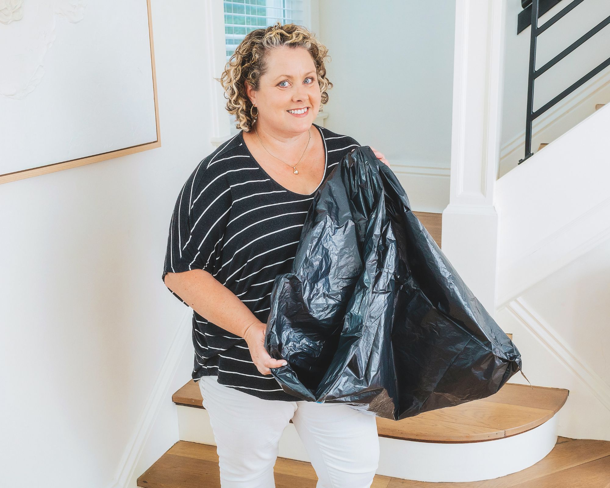Dana White is a white woman with curly blonde hair, pictured in a black mid-sleeve t-shirt with white stripes. She is in a minimalist hallways, holding a black trash bag with stairs in the background