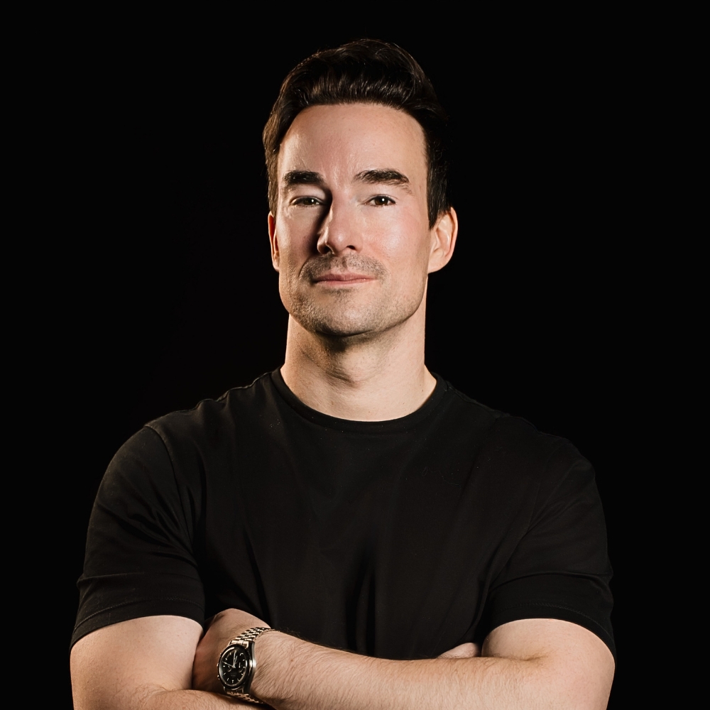 Man with dark hair and crossed arms, wearing a black t-shirt, studio portrait on a black background.