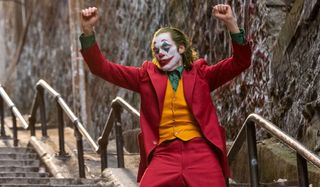 the Joker dancing on the stairs in a red suit with orange vest and green shirt.