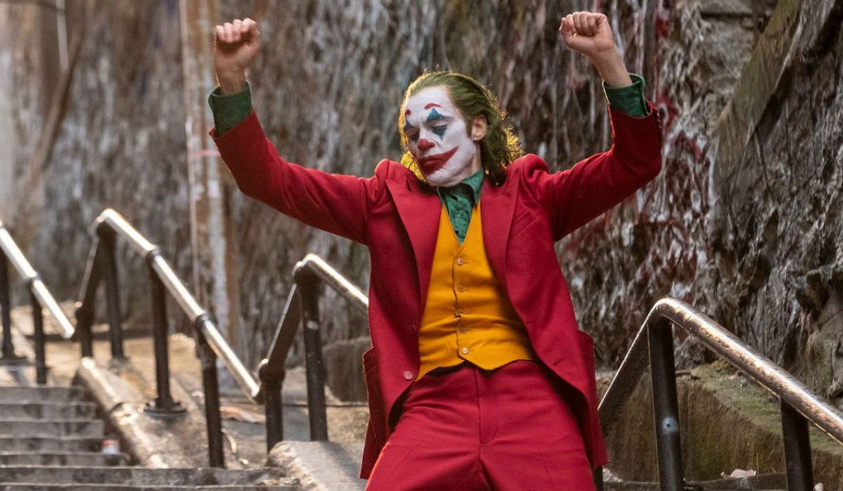 the Joker dancing on the stairs in a red suit with orange vest and green shirt.