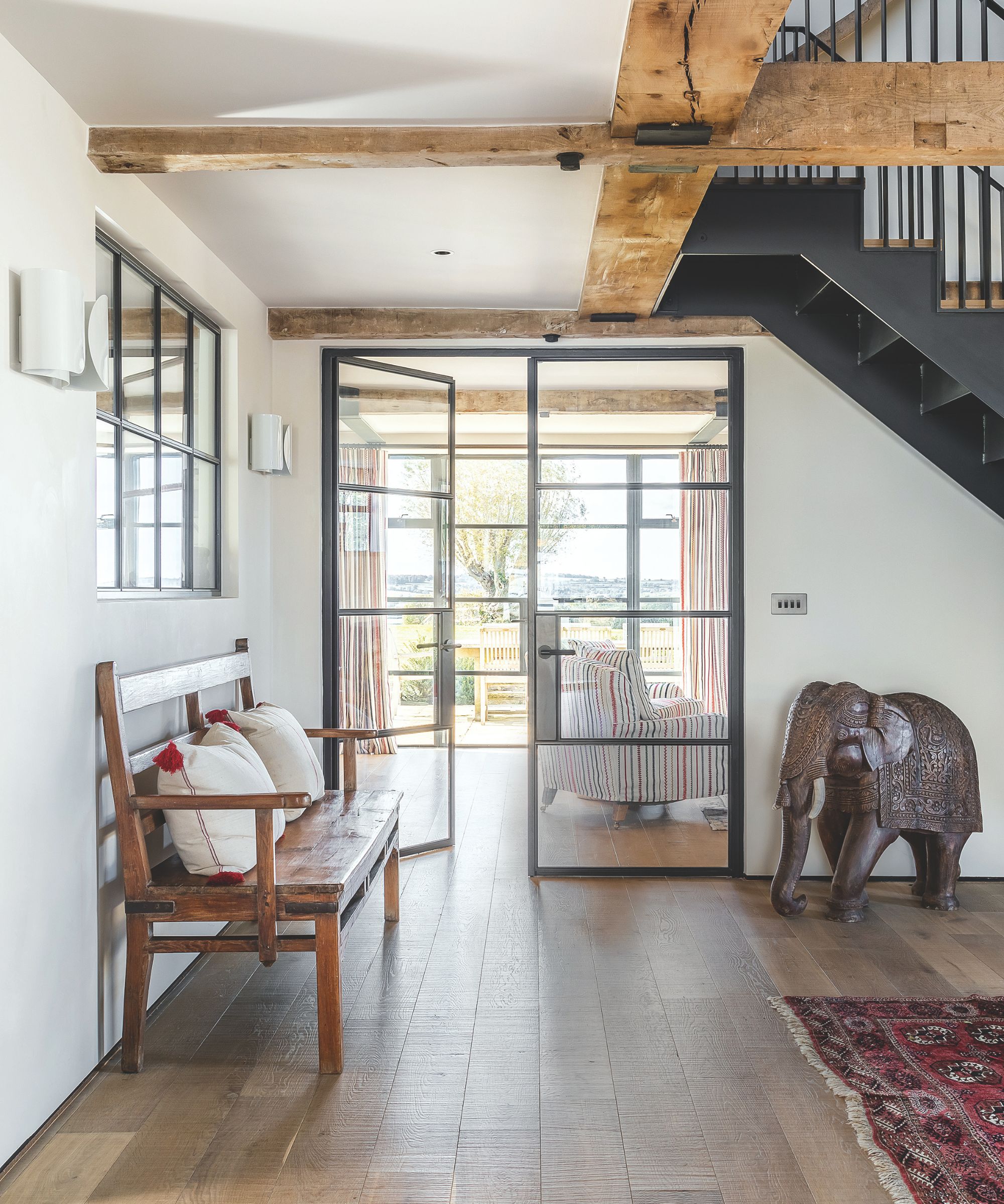 Hallway with wooden beams, metal staircase, wooden floor with red Persian rug, antique elephant statue and wooden bench