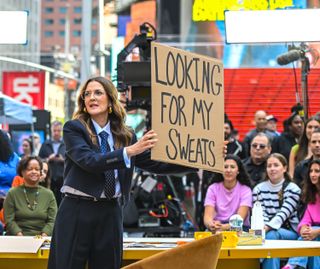 Drew Barrymore wears a suit and tie and holds up a sign that says looking for my sweats