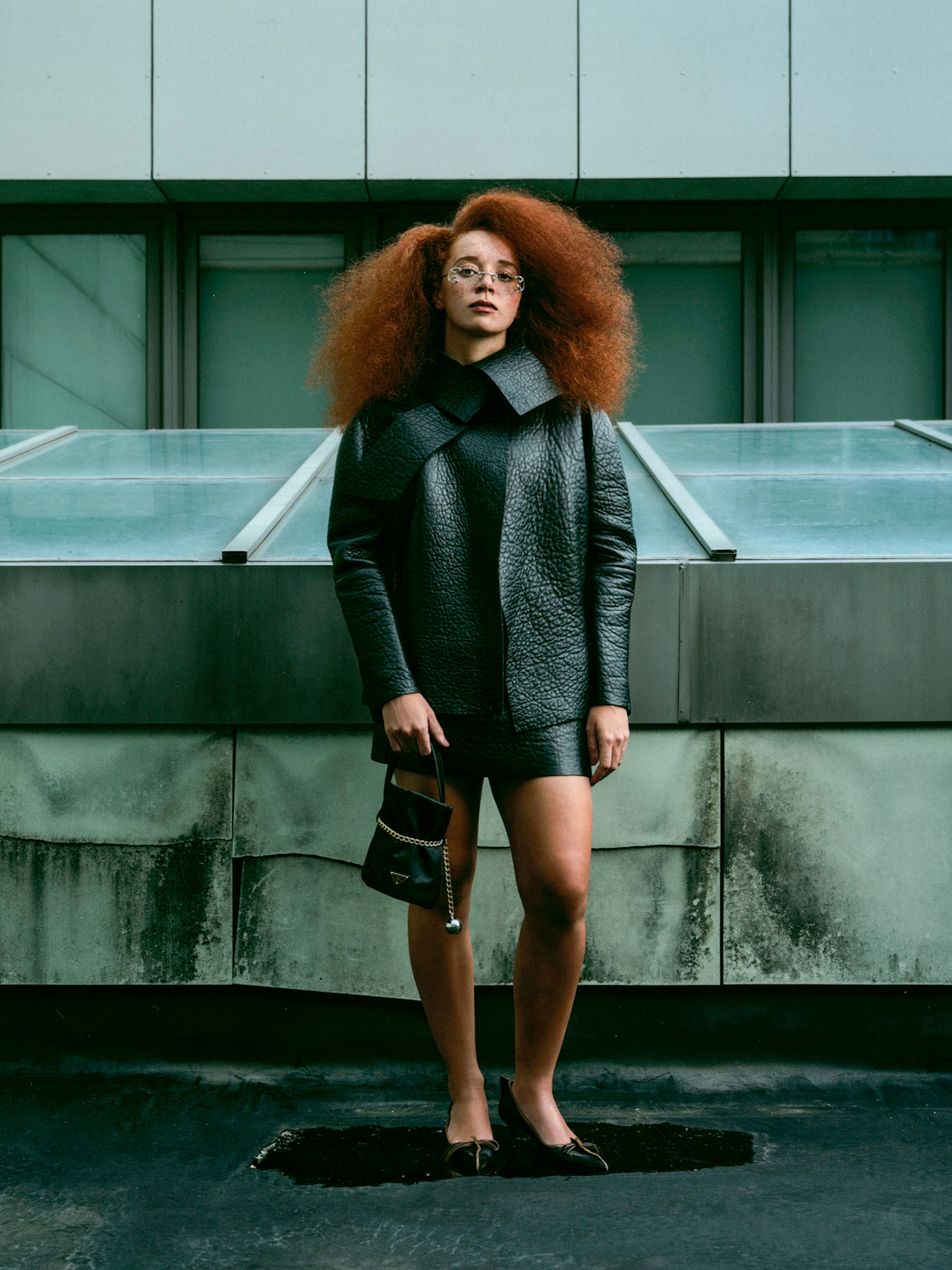 Photo of Erin Kellyman for the Fall Ones to Watch portfolio. She is pictured against an urban backdrop wearing a pebbled Prada leather jacket, metal-accented glasses, pointed-toe pumps, and a Prada bag.