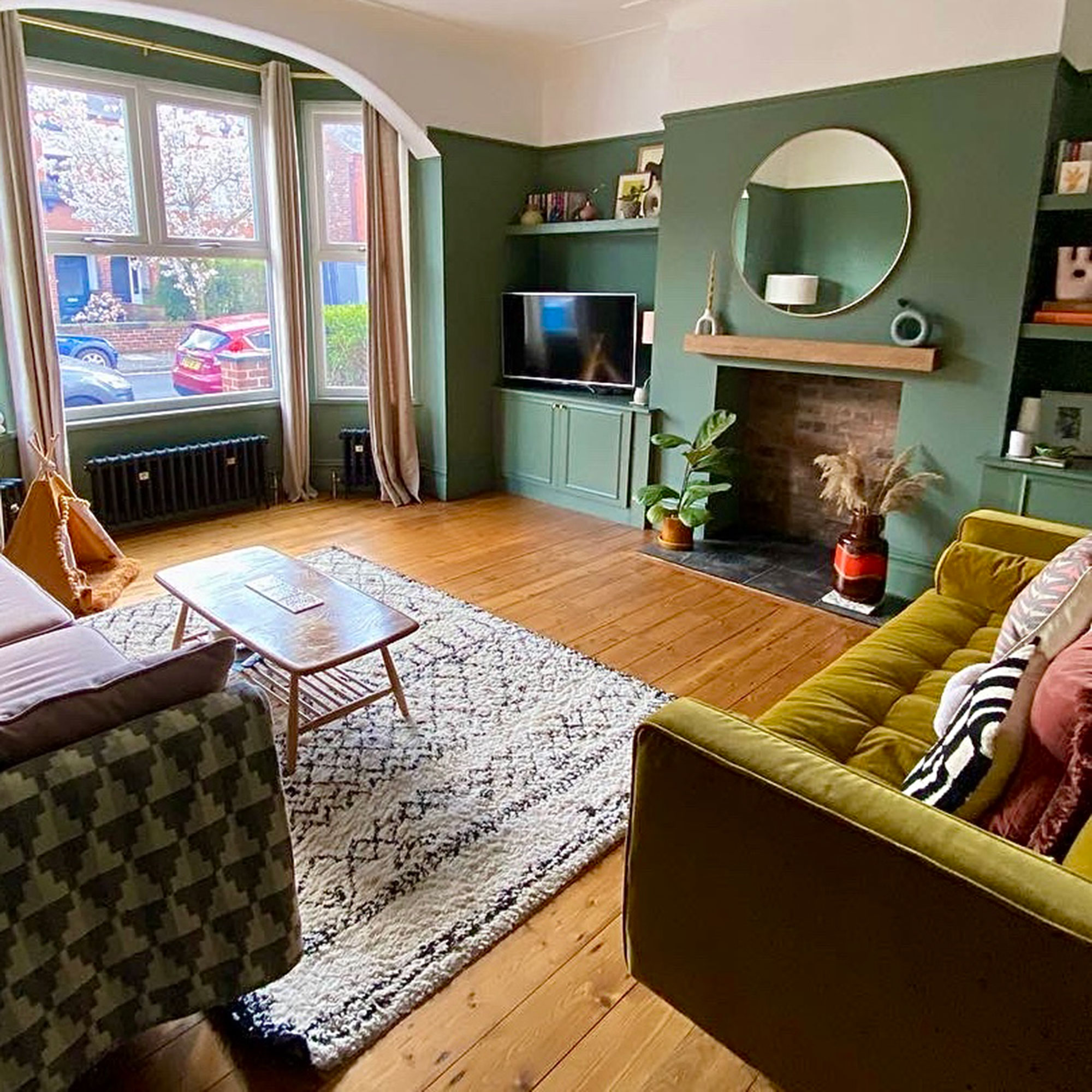 Green living room with wooden floor, Berber rug, green velvet sofa and large round mirror over fireplace alcove