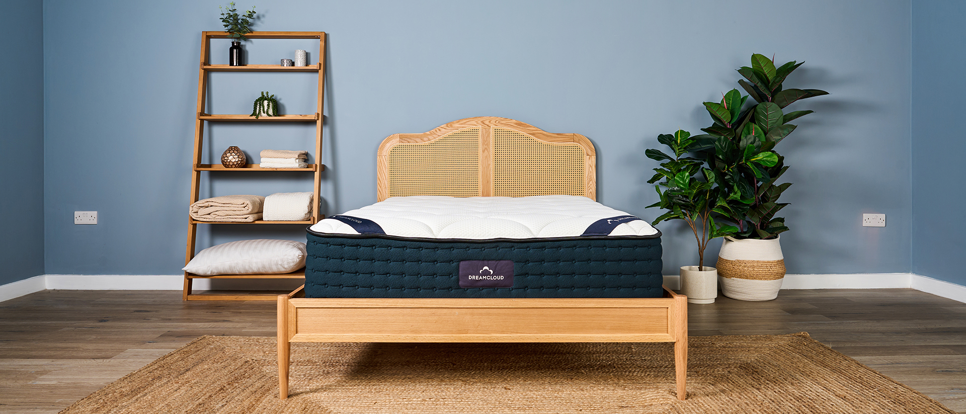 The DreamCloud Hybrid Mattress on a wooden bed frame in our testing studio, ready to be used by our review team. To the left of the bed is a set of shelves, to the right are two potted plants