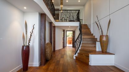 Large, modern entryway with wooden staircase and wooden floors, and large ornate vases. Second floor is partially pictured behind black banister.