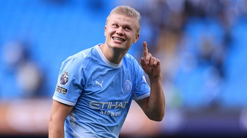Erling Haaland, wearing Manchester City blue, makes a No1 signal while interacting with fans.