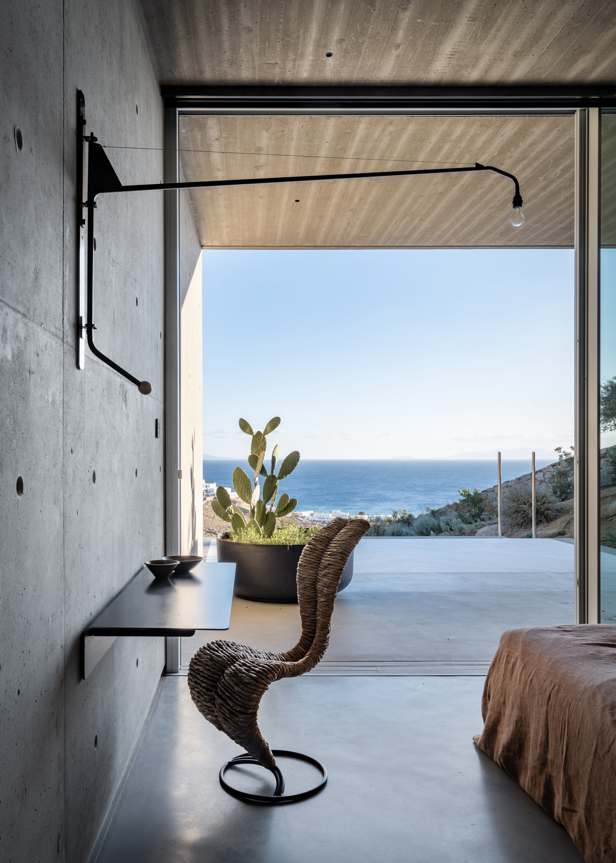 coastal brutalist interior with concrete walls and floor, swivel light mounted on wall, floating desk with tom dixon chair, with edge of bed