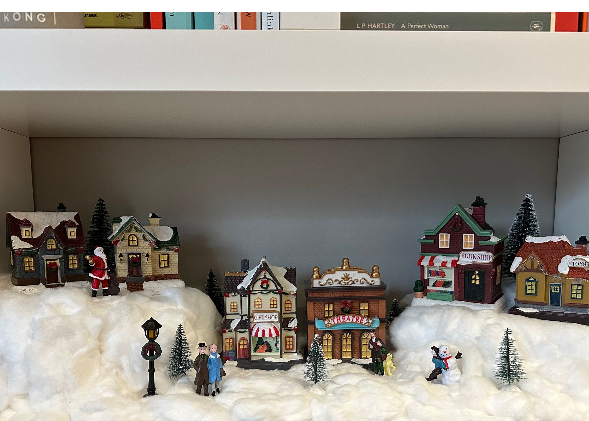 A miniature christmas village in a book cubby with fake snow, houses on multiple levels, and trees, figures and a lampost just off the center