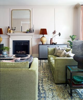 Pale pink painted living room with green and blue rug, green sofas, fireplace, black coffee table