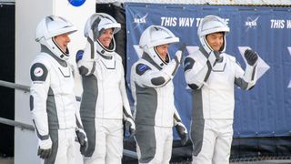 four astronauts in white spacesuits stand and wave outside a nasa building