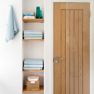 Small alcove in a bathroom with bespoke brown shelves holding blue spotted towels, white walls and brown door