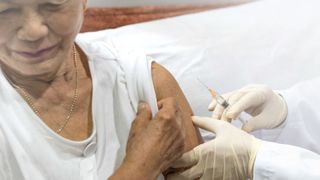 older woman with light grey hair and tan skin raises the arm of her t shirt to receive a shot from a gloved medical provider who's holding a syringe at the ready