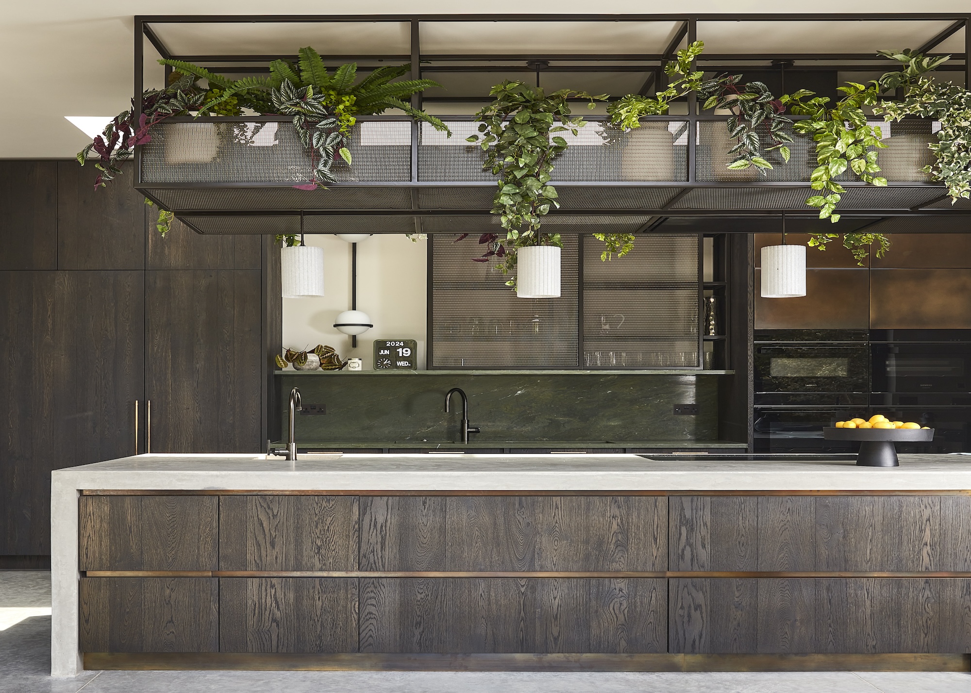 A contemporary kitchen in dark stained oak showing a mix of handle-free cabinets on the island and bar handles on the tall, wall units, also shown with a ceiling storage feature filled with plants