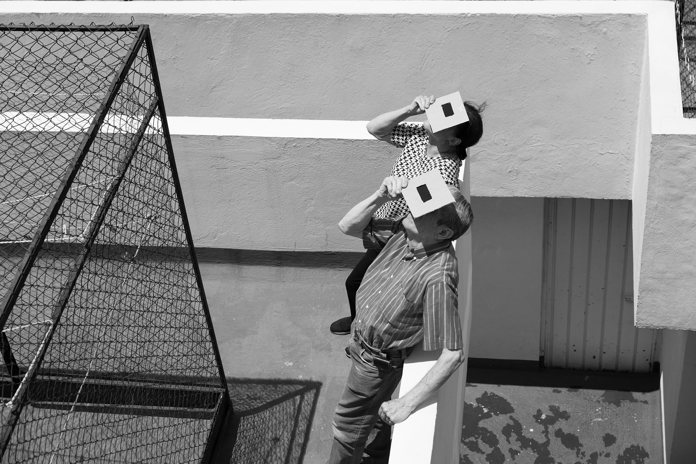 Two people, facing up, cover their heads with cardboard boxes having square cutouts. The setting seems outdoors with geometric shadows and lines