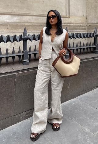 a woman's outfit showing what to wear in 80-degree weather with a tan linen vest with matching trousers, flatform sandals, and a tan tote bag