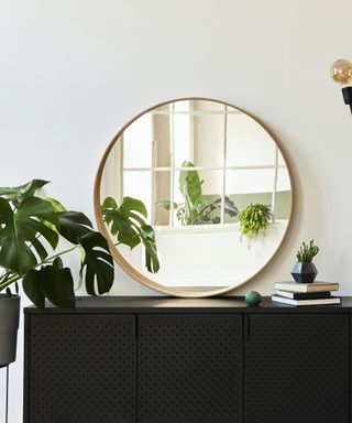 A black storage console with a large gold round mirror on it and a plant next to it