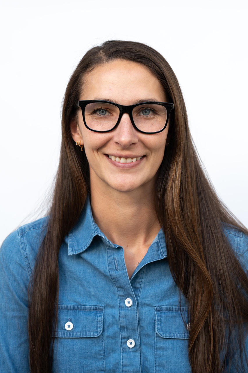 Woman smiling with long brown hair in blue shirt and black glasses