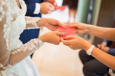 A person hands a red envelope to a woman wearing a long-sleeved wedding gown. 
