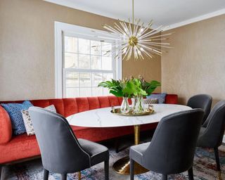 Rich rust banquette style seating and upholstered slate gray dining chairs around oval marble and brass pedestal dining table, with starburst feature pendant.