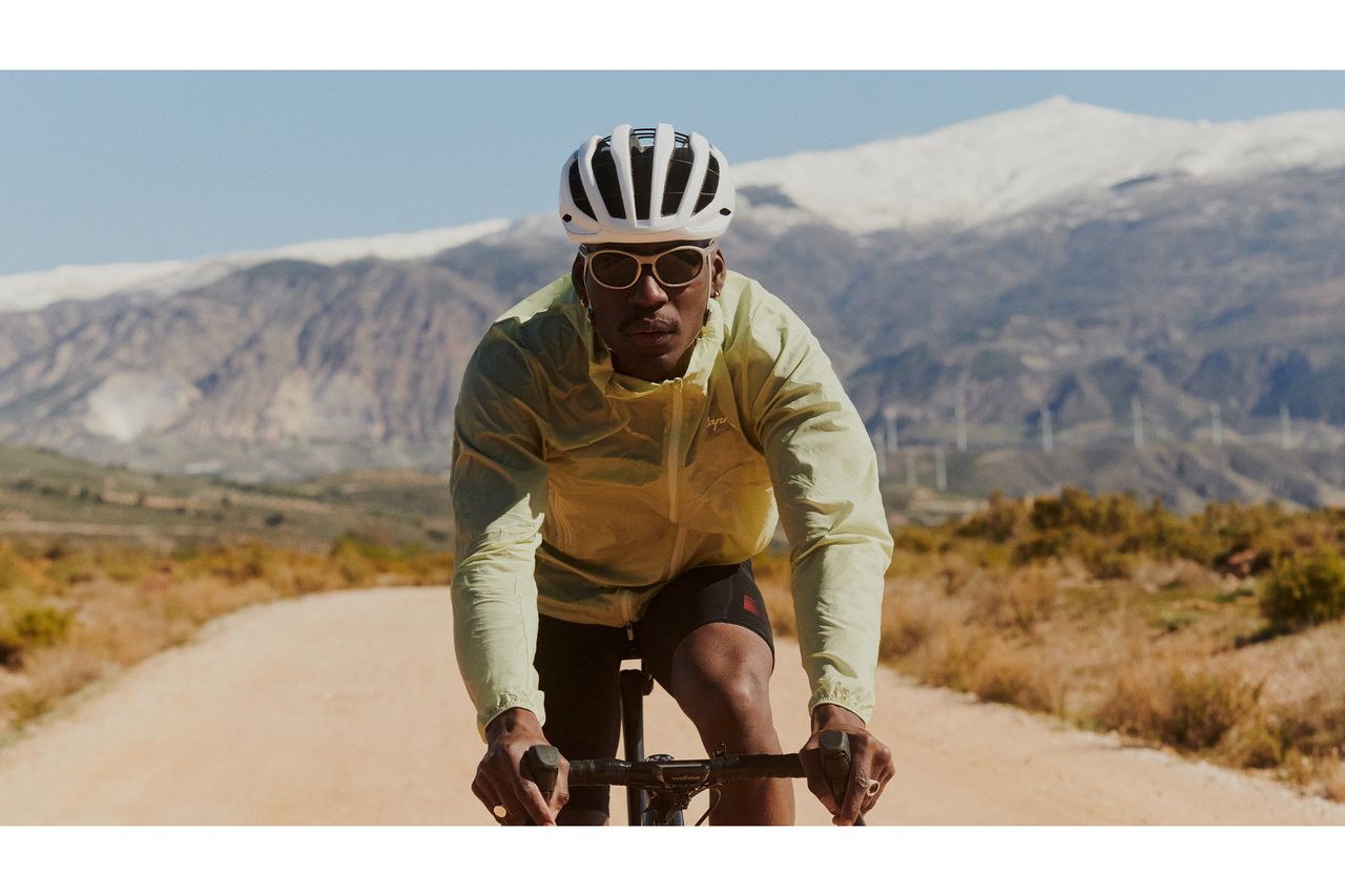 A man in a yellow windbreaker and white helmet rides towards the camera wearing the Rapha Dalton sunglasses