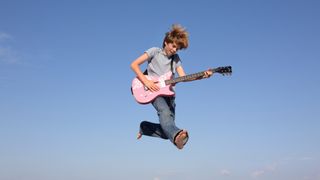 Boy playing pink guitar jumps in the air against a blue sky