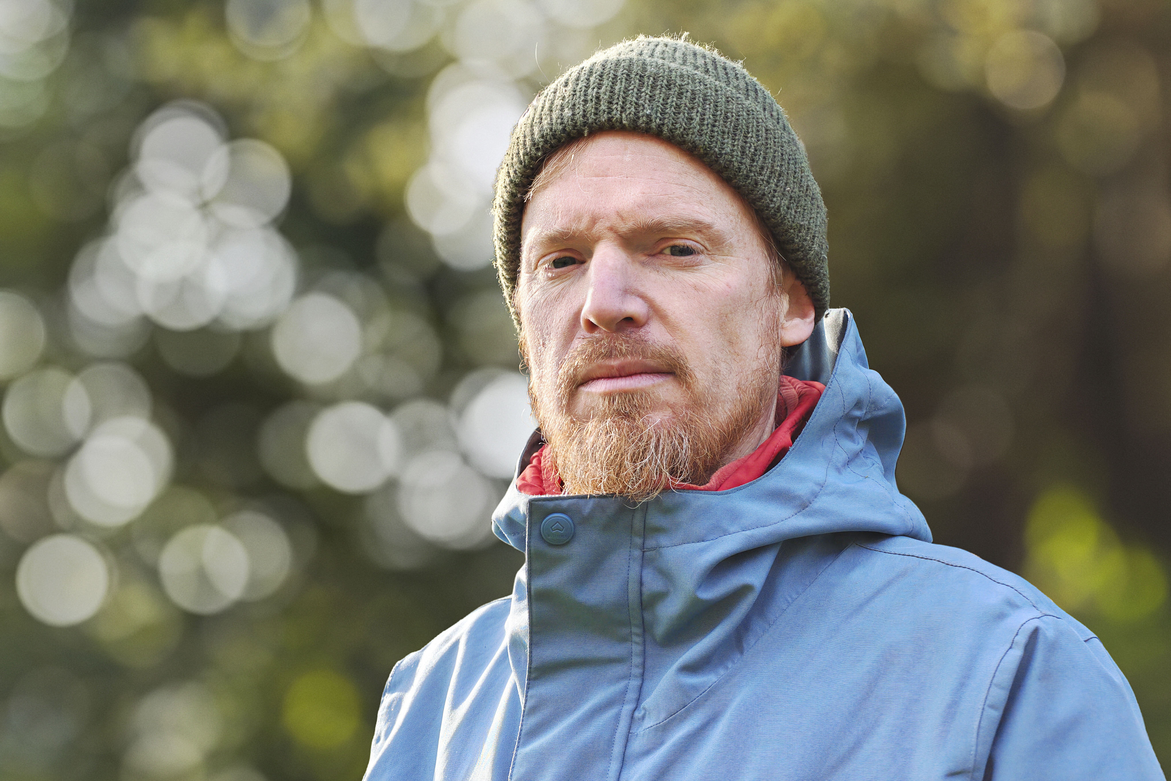 Portrait of a man in a blue coat and green beanie, with dappled light through a background tree