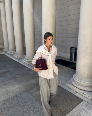 A woman wears a stripe button-down shirt, white sweater, red Bottega Veneta Andiamo bag, and gray trousers.