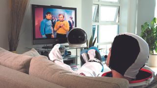 person dressed as an astronaut takes a lie down on the couch and watches television.