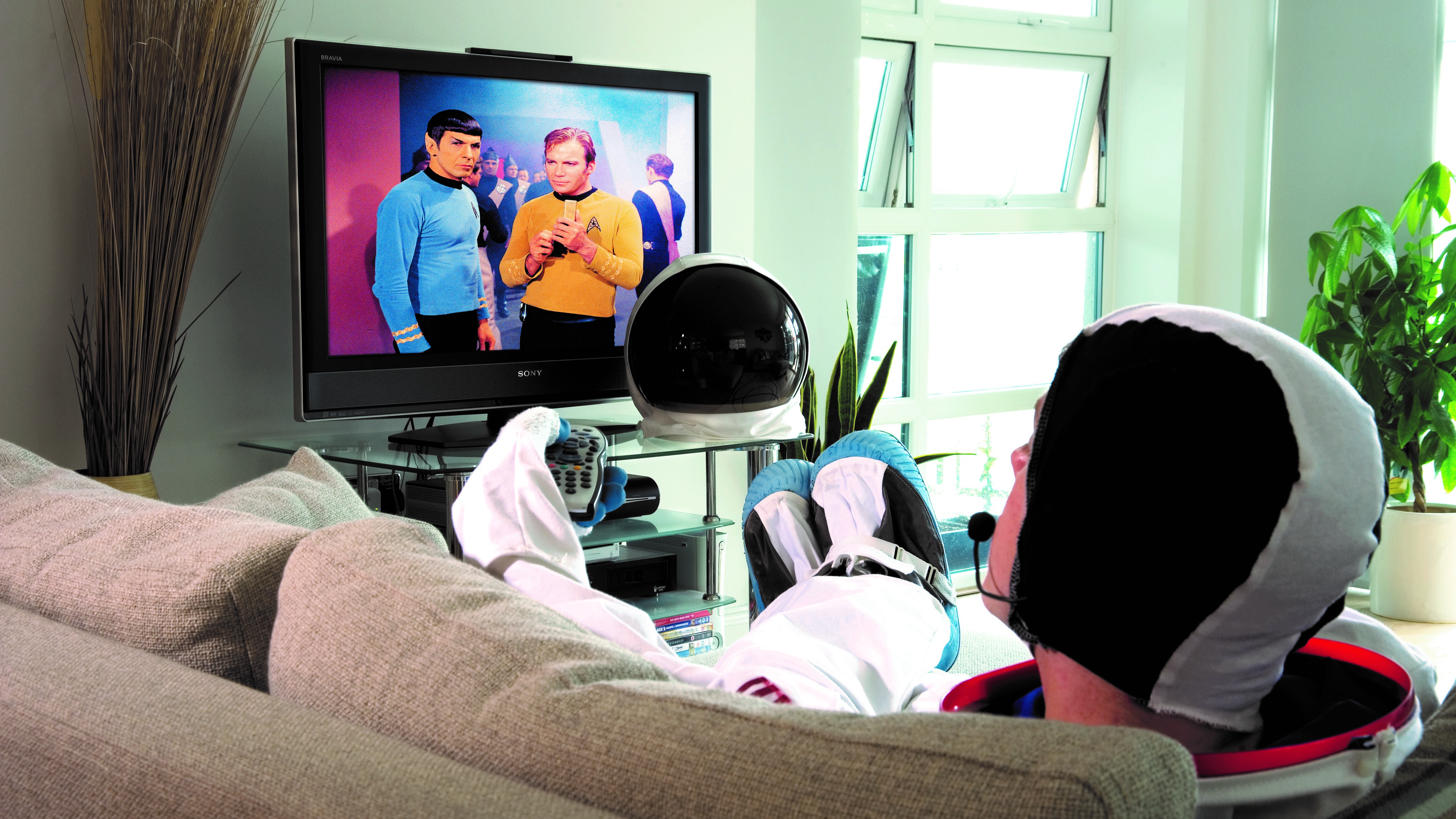 A person dressed as an astronaut is lying on the sofa and watching TV.