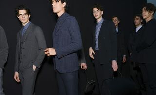 Six male models wearing looks from Giorgio Armani's collection. They are wearing blue and grey suits, shirts and waistcoats. One model is holding a bag and there is a seventh person hidden behind one of the models also holding a bag